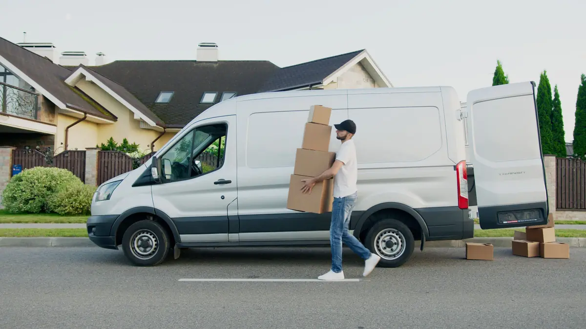 A delivery driver carries a stack of boxes beside a white cargo van with its rear door open.